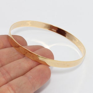 May include: A gold-colored bangle bracelet. The bracelet is a simple, smooth band with a polished finish. The jewelry is round and appears to be made of metal. The bracelet is held in a hand against a white background.