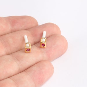 May include: A pair of small, gold-colored earrings shaped like nail polish bottles. Each earring features a white brush, a gold bottle, and a small, round, red gemstone. The earrings are displayed on a light background.