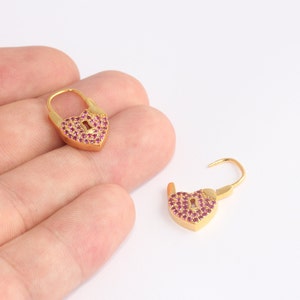 May include: Two gold-colored heart-shaped earrings. Each earring is a padlock design, with a keyhole detail. The heart shape is encrusted with small, round, red gemstones. One earring is open, and the other is closed.