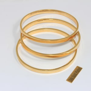 May include: Three smooth, polished gold-tone bangle bracelets stacked together. Each bangle has a simple, rounded profile with a reflective finish. A small rectangular tag placed near the bracelets features the text 24k Gold Plated.