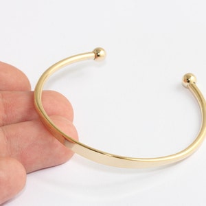 May include: A gold-coloured open cuff bracelet with a smooth, polished finish. The bracelet has a rounded shape with a small ball at each open end. The jewellery is shown against a white background.