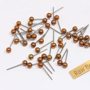 May include: A collection of raw brass ball-end pins with silver-colored stems, scattered on a white surface. A small, rectangular brass tag with the text "Raw Brass" is also present. The pins have a copper-colored finish.
