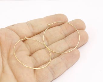 Puede incluir: Un par de pendientes de aro dorados. Los pendientes son finos y circulares, con un diseño simple y minimalista. Los aros se sostienen en una mano sobre un fondo blanco. Los pendientes son probablemente de metal.