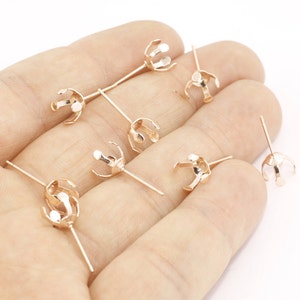 May include: Several rose gold-colored jewelry findings are displayed. Each finding has a pronged setting at the top, designed to hold a gemstone, and a long, slender post. The findings are scattered across a light-colored surface.