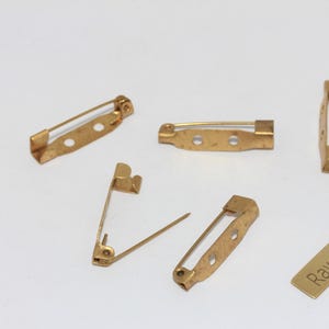 May include: Several small, gold-colored brass pin backs are scattered on a white surface. One pin back has a needle extended. A small rectangular brass tag with the words "Raw Brass" is also present. These are likely used for jewelry making or crafts.