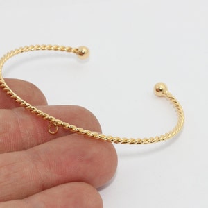 Può includere: Un bracciale aperto color oro con un design a corda intrecciata. Il bracciale ha una piccola sfera a ciascuna estremità e un piccolo anello per attaccare i ciondoli. Il gioiello è mostrato su sfondo bianco.