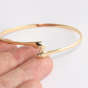 以下が含まれることがあります： 開いた端を持つ金色のバングルブレスレットで、触れ合おうとする2つの小さな手が付いています。ブレスレットは光沢のある滑らかな金属製で、白い背景に配置されています。