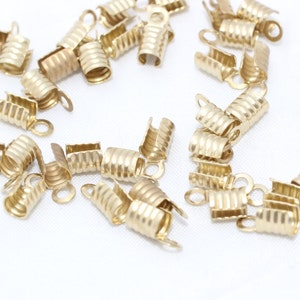 Puede incluir: Una colección de pequeños terminales de crimpado dorados para la fabricación de joyas. Cada pieza tiene un diseño acanalado y un pequeño lazo para la fijación. Los terminales están dispersos sobre una superficie blanca.