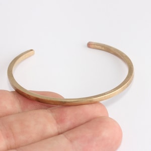 May include: A gold-colored cuff bracelet with a simple, open design. The bracelet has a slightly hammered texture and a rounded rectangular shape. The jewelry is shown on a white background.