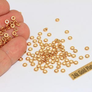 May include: A collection of small, round, gold-colored metal spacer beads with a central hole. A rectangular gold-colored metal tag is present with the text 24k Gold Plated printed in black. The beads are scattered and held in a hand.