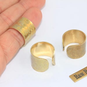 Könnte beinhalten: Drei Messingringe, zwei offen und einer an einem Finger getragen, mit gehämmertem Muster. Ein kleines Messingrechteck mit dem Text "Raw Brass" ist ebenfalls vorhanden. Die Ringe haben eine warme Goldfarbe.