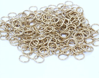 May include: A pile of small, gold-colored metal rings. The rings are round and appear to be made of a shiny, possibly brass or gold-plated material. They are scattered across a white surface.