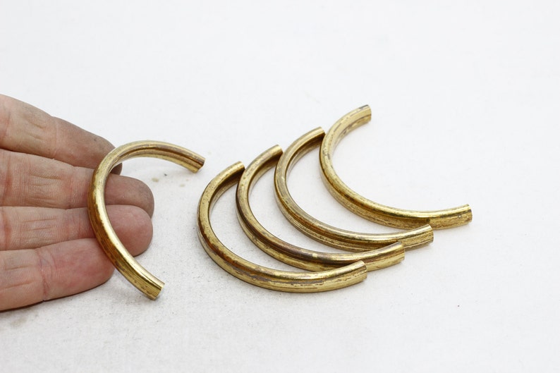 May include: A collection of curved, brass-colored metal pieces. The pieces are semi-circular, with a rounded profile and a slightly textured surface. The objects are arranged on a white surface, with one piece held by a hand.