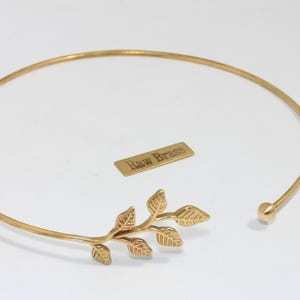 Könnte beinhalten: Ein goldfarbenes Choker-Halskette mit Blattdesign. Die Halskette hat ein zartes, offenes Design mit einem kleinen Ball am Verschluss. Ein kleines rechteckiges Etikett mit dem Text "Raw Brass" ist ebenfalls vorhanden.