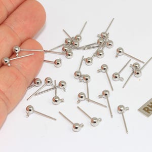 May include: A collection of silver-toned metal ball stud earring findings with small loops for attaching charms. Each piece features a smooth spherical top and a straight post. A small rectangular metal tag with the word RHODIUM is included.