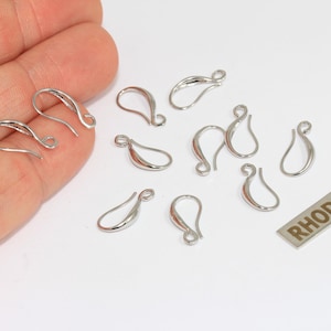 May include: A collection of silver-toned metal earring hooks with a curved, teardrop-shaped design. The set includes ten individual earring findings and a small rectangular metal tag with the word RHODIUM printed in dark capital letters.