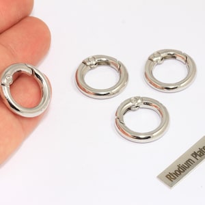 May include: Silver-colored round spring clasp findings. One clasp is held in a hand, with three others and a small metal tag that reads "Rhodium Plated" on a white background. These clasps are used for jewelry making.