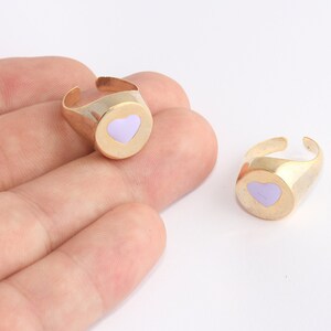 Puede incluir: Dos anillos ajustables dorados con un corazón de esmalte lavanda. Los anillos tienen un diseño sencillo y moderno y se muestran sobre un fondo blanco.
