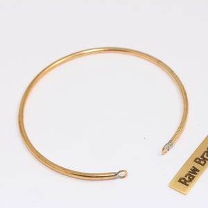 Pode incluir: Uma pulseira aberta dourada com um design circular. A pulseira é feita de um metal fino e liso. Uma pequena etiqueta retangular com as palavras "Raw Brass" está ao lado da pulseira.