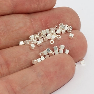 Könnte beinhalten: Eine Ansammlung kleiner, silberfarbener Würfelperlen. Die Perlen sind auf einer Oberfläche verstreut, einige werden in einer Hand gehalten. Jede Perle hat ein kleines Loch in der Mitte, was darauf hindeutet, dass sie für die Schmuckherstellung oder für Bastelarbeiten gedacht sind.