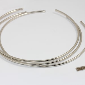 Könnte beinhalten: Mehrere silberfarbene, offene Choker-Halsketten sind auf einer weißen Oberfläche angeordnet. Ein kleines rechteckiges Etikett mit dem Text "Rhodium Plated" ist ebenfalls vorhanden. Die Halsketten bestehen aus glänzendem Metall.