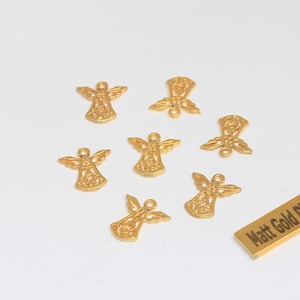 以下が含まれることがあります： 金色の天使チャーム。翼と体のデザインが施されています。白い表面に散らばっており、「Matt Gold Plated」と書かれた長方形のタグが付いています。ジュエリー作りやクラフトに最適です。