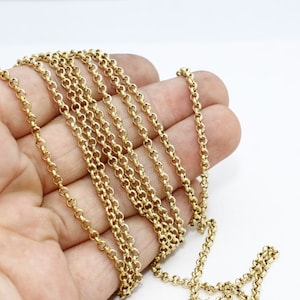 May include: A gold-colored chain necklace with a rounded link design. The chain is draped across a hand, showcasing its length and detail against a white background. The chain's links are closely connected, creating a continuous pattern.
