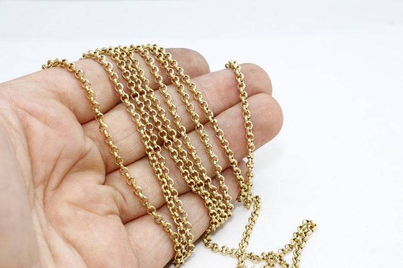 May include: A gold-colored chain necklace with a rounded link design. The chain is draped across a hand, showcasing its length and detail against a white background. The chain's links are closely connected, creating a continuous pattern.