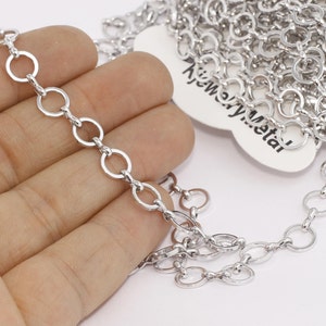 May include: A roll of silver-colored chain with a series of small, connected circular links. The chain is displayed on a white background, with a portion of the chain extending towards the viewer. The chain is made of metal.