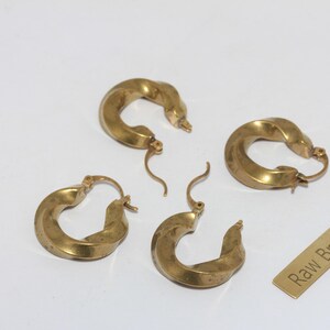 Op de afbeelding: Vier goudkleurige messing oorringen met een gedraaid ontwerp. De oorbellen zijn gerangschikt op een wit oppervlak, met een klein rechthoekig label met de tekst "Raw Brass".