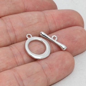 Könnte beinhalten: Ein silberfarbener Knebelverschluss, bestehend aus einem kreisförmigen Ring und einem Stab mit einer Schlaufe. Dieser Schmuckherstellungsbestandteil wird vor einem neutralen Hintergrund gezeigt.