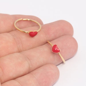 Pode incluir: Dois anéis dourados com um pequeno pingente em forma de coração vermelho. Os anéis são finos e delicados, com um acabamento polido. Os pingentes de coração são de cor vermelha vibrante, adicionando um toque de cor ao design minimalista. Em fundo branco.