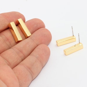 Könnte beinhalten: Zwei Paar goldfarbene Ohrstecker in Stäbchenform. Ein Paar wird in einer Hand gehalten, während das andere Paar auf einer weißen Oberfläche liegt. Die Ohrringe haben eine rechteckige Form mit einem Stecker zum Durchstechen.
