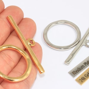 May include: Close-up of jewelry findings, including gold and silver toggle clasps. One gold clasp is in a hand. Two small rectangular tags read "Rhodium Plated" and "24k Gold Plated".
