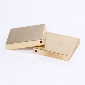 Puede incluir: Dos componentes metálicos rectangulares de color dorado, cada uno con un pequeño agujero perforado. Los objetos están colocados sobre un fondo blanco. Los artículos parecen ser elementos de ferretería o decorativos.