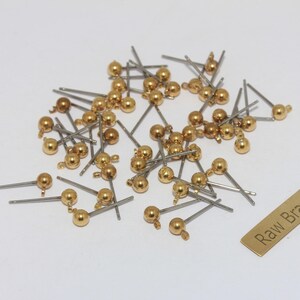 Può includere: Una collezione di piccoli componenti a forma di sfera color oro con perni color argento, sparsi su una superficie bianca. È presente anche una piccola etichetta rettangolare in ottone con la scritta "Raw Brass". Questi componenti sembrano essere componenti per gioielli.