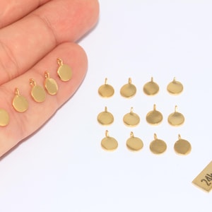 May include: Several small, gold-coloured, round pendants with a loop at the top, ideal for jewellery making. Some are held in a hand, others are on a white surface. A small rectangular tag reads "24k Gold Plated".