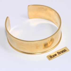 Puede incluir: Un brazalete de puño de latón ancho y abierto con un acabado cepillado. Una pequeña etiqueta de latón junto al brazalete dice "Raw Brass". El brazalete es de color dorado cálido.