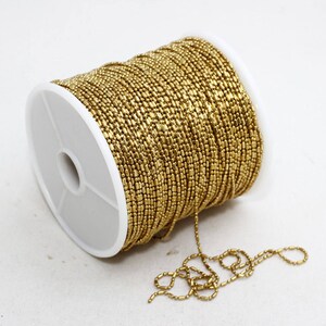 May include: A spool of gold-colored beaded chain. The chain is tightly wound around a white plastic spool. A length of the chain extends from the spool, lying on the white surface. The beads are small and rectangular.