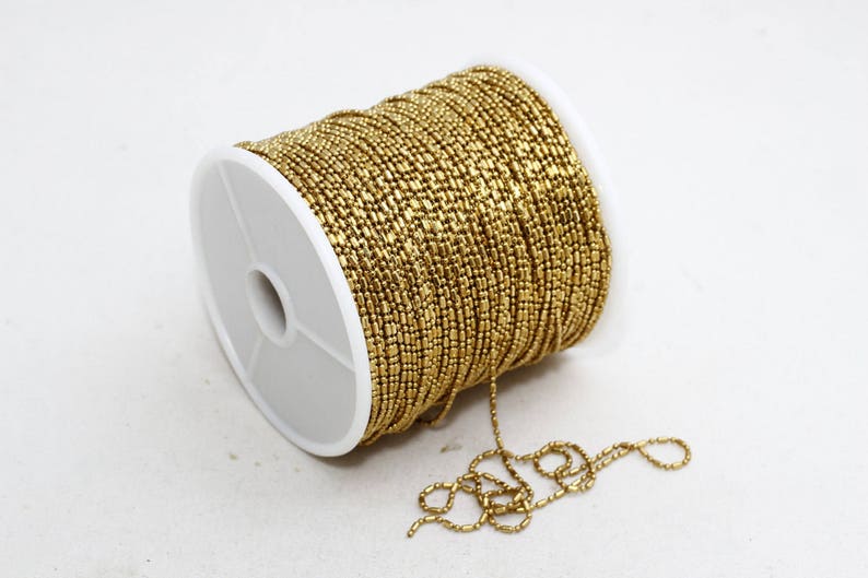 May include: A spool of gold-colored beaded chain. The chain is tightly wound around a white plastic spool. A length of the chain extends from the spool, lying on the white surface. The beads are small and rectangular.