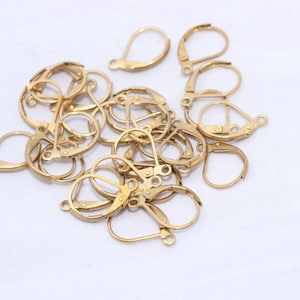May include: A selection of gold-coloured earring hooks, or leverback earring findings, are scattered on a white surface. The metal earring components feature a hinged design for secure closure. These findings are used for jewellery making.