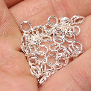 May include: A handful of small, silver jump rings, displayed in an open palm. The rings are circular, with a small gap for connecting to other jewellery elements. The image is well-lit, showing the metallic shine of the rings.