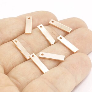 Peut inclure: Plusieurs petits breloques rectangulaires en métal de couleur or rose, avec un seul trou en haut. Les breloques sont dans une main, et sont probablement utilisées pour la fabrication de bijoux ou d'autres travaux manuels.
