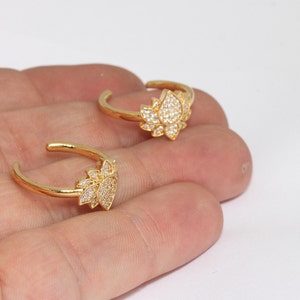 Puede incluir: Dos anillos ajustables dorados, cada uno con un diseño de flor de loto. La flor de loto está incrustada con pequeñas piedras preciosas transparentes. Los anillos se muestran sobre un fondo neutro.