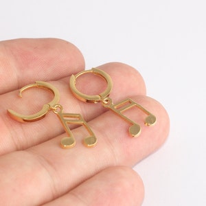 Puede incluir: Pendientes dorados con forma de nota musical. Cada pendiente presenta un pequeño aro con un dije de nota musical colgante. Los pendientes se muestran sobre un fondo claro, resaltando su acabado metálico brillante. Un accesorio elegante para amantes de la música.