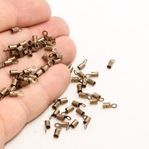 Puede incluir: Pequeños terminales de crimpado de color latón antiguo con un lazo para la fabricación de joyas. Estos componentes metálicos son cilíndricos con un pequeño lazo en un extremo, perfectos para sujetar a cordones o cintas.