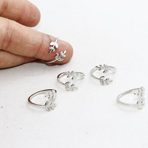 May include: Silver-toned leaf rings. The rings feature a delicate leaf design and are adjustable. One ring is worn on a finger, with several others displayed on a white surface. The rings are likely made of metal.