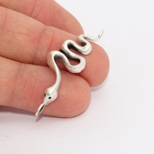 Peut inclure: Un charm en forme de serpent argenté avec un corps incurvé et une petite tête. Le serpent a des yeux en forme de points noirs et une bouche légèrement ouverte. Le charme est sur fond blanc.