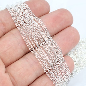 May include: Close-up of several strands of delicate, silver-coloured chain. The chains are made of small, interconnected oval links. The chains are held in a hand against a white background. The chains are likely for jewellery making.