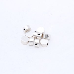 Può includere: Una collezione di piccole perline cilindriche color argento con una parte superiore e inferiore piatte. Ogni perlina ha un piccolo foro che attraversa il centro. Le perline sono sparse su uno sfondo bianco.
